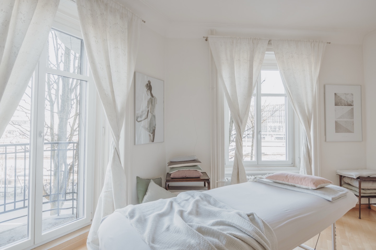 Behandlungsraum für Craniosacral Therapie in der Praxis Zentral an der Zentralstrasse 38 in Luzern: Massageliege mit weisser Decke vor hohen Altbaufenstern mit hellen Vorhängen.